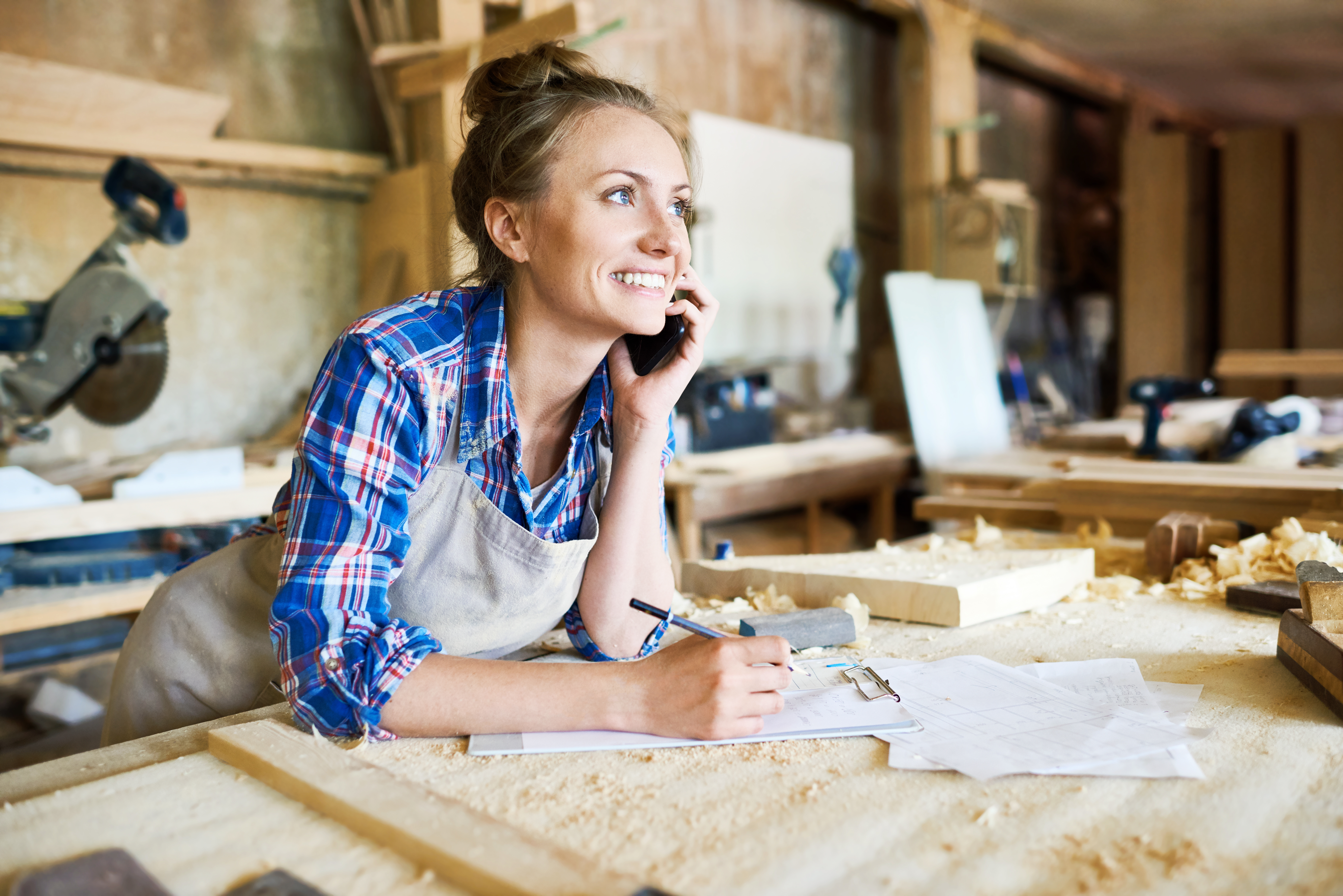 Cheerful young carpenter discussing order details with her customer on smartphone and taking necessary notes, interior of spacious workshop on background